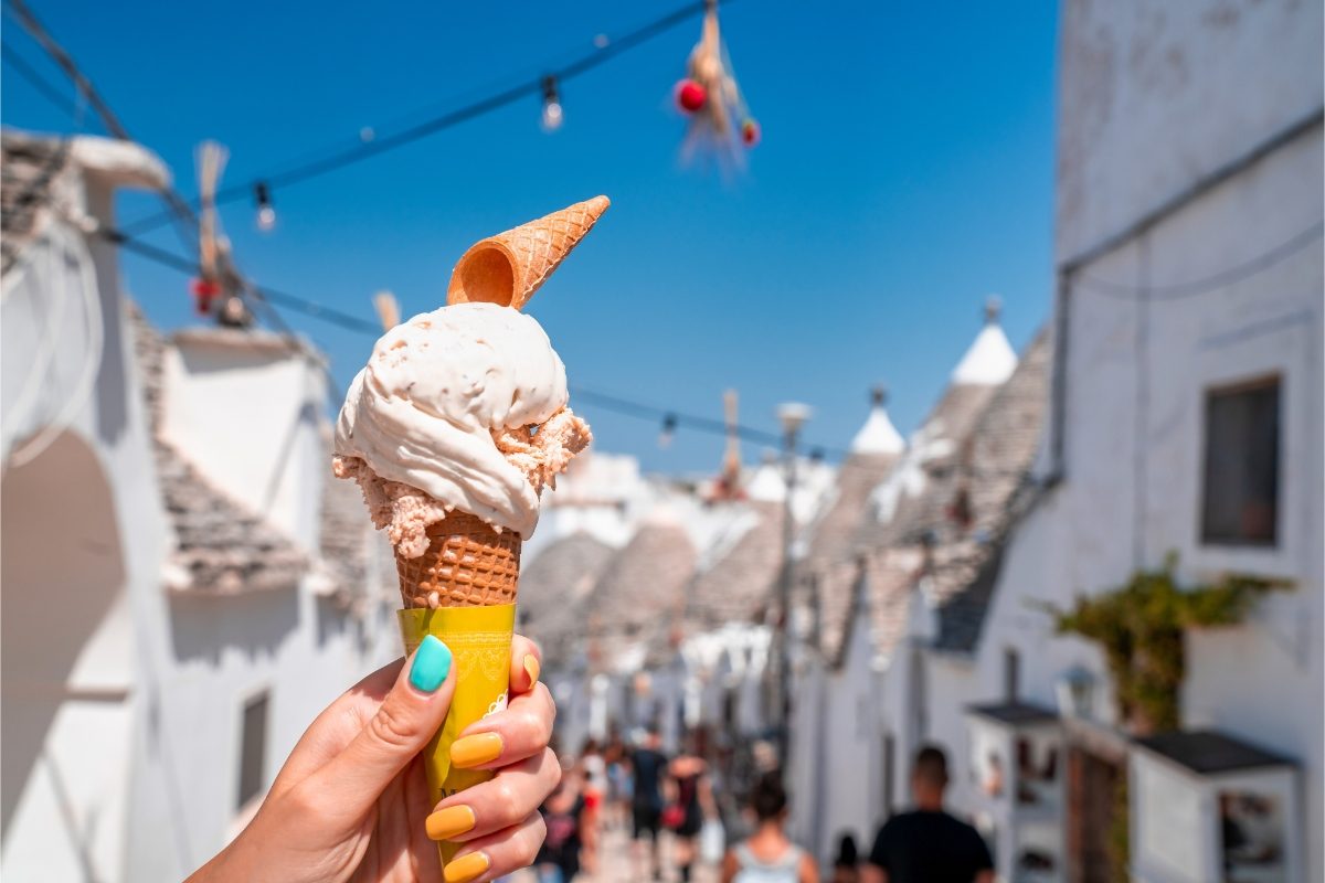 Close-up of a hand holding a gelato with trulli houses in Alberobello in the background, in Bari, Puglia, Italy