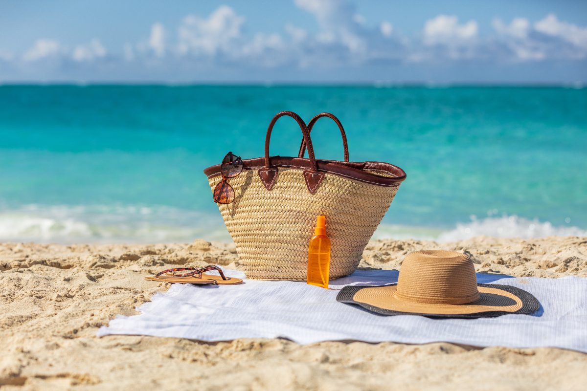 Beach bag sits next to sunglasses, sunscreen, sandals, and a wide-brimmed hat