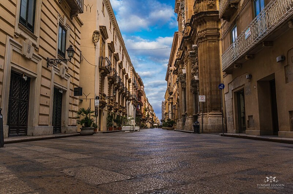 A charming street view in Italy, lined with colorful buildings and lush greenery, leading to a quaint vacation accommodation nestled among the picturesque surroundings.