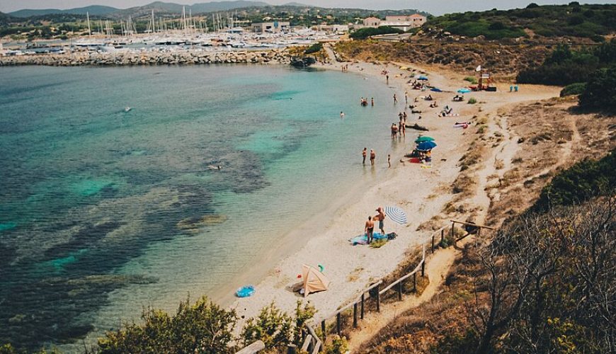 A serene beach in Italy with golden sands, crystal-clear turquoise waters, and distant hills under a clear blue sky.