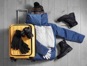 Top view of winter clothes, boots, a camera, and a yellow suitcase on a wooden floor