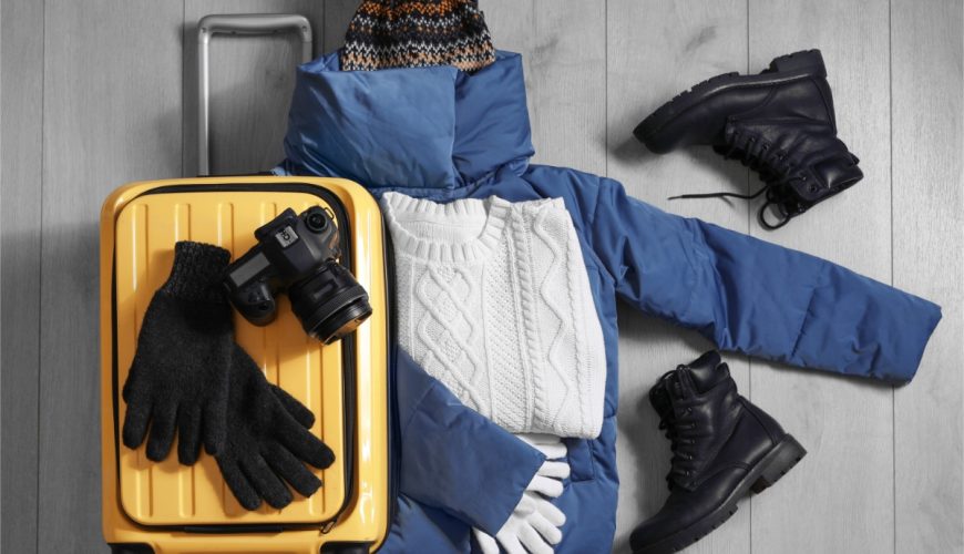 Top view of winter clothes, boots, a camera, and a yellow suitcase on a wooden floor