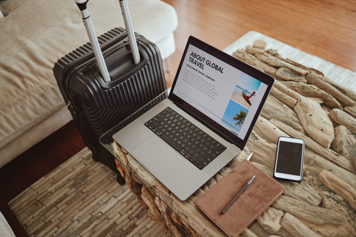 Black luggage sits next to a laptop displaying global travel plans, accompanied by a phone and a travel journal on the table