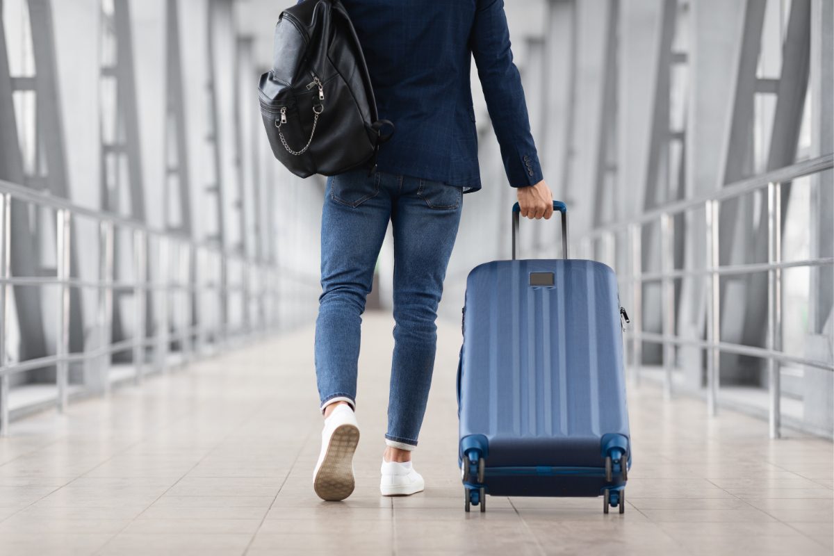 Man with a bag slung over his shoulder and a suitcase in hand walks through an airport